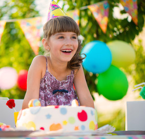 Little girl smiling, sitting in front of a cake and wearing a birthday hat, with balloons in background